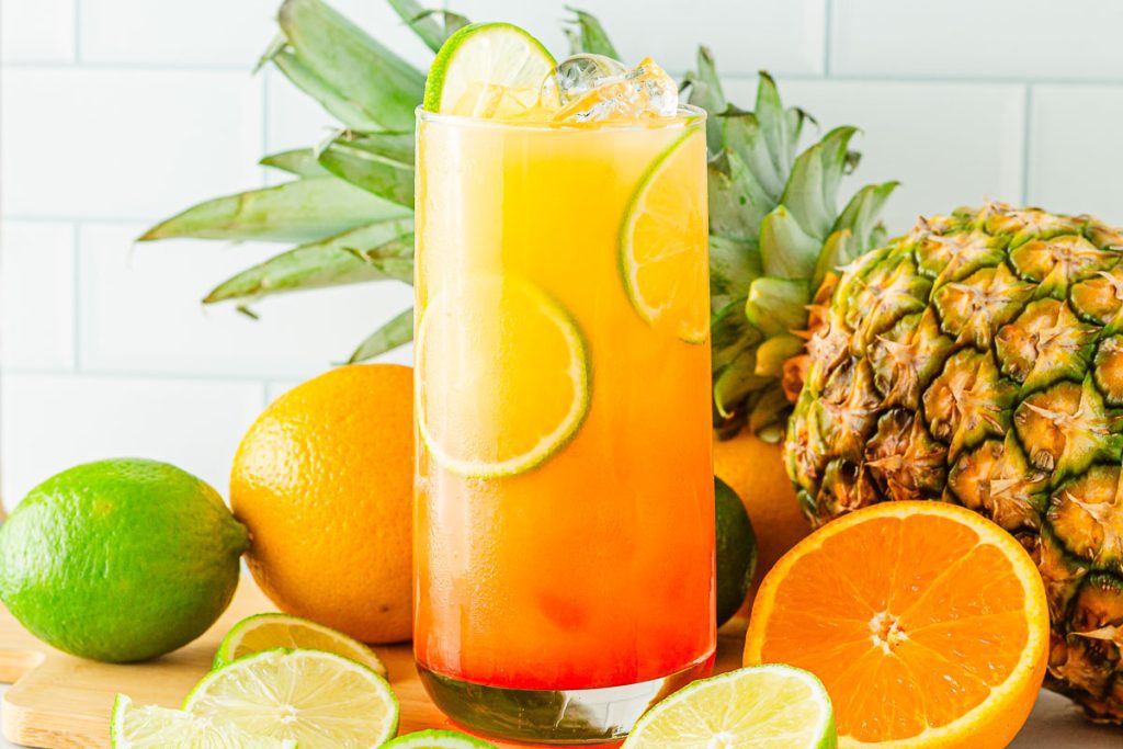 glass of non-alcoholic Virgin Tequila Sunrise Mocktail with limes and oranges and a pineapple