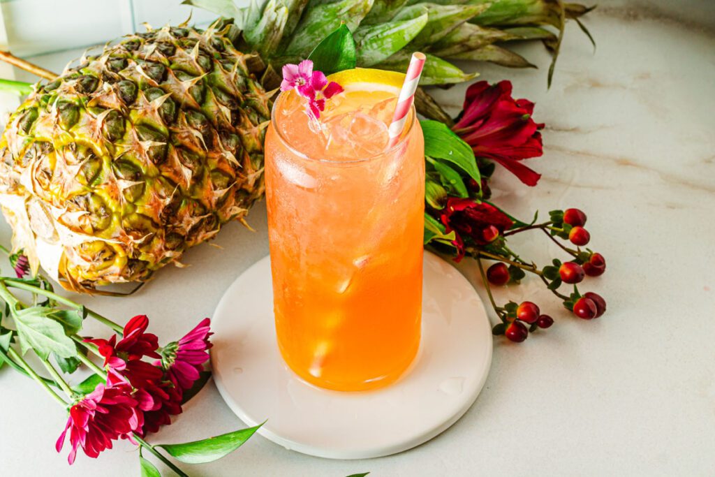glass of pineapple tropical mocktail surrounded by flowers and a pineapple